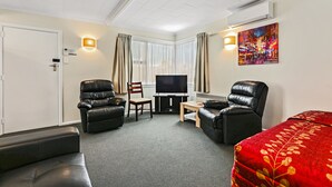 A hotel room with a large bed, two black leather sofas, a wooden chair, a small table, a flat-screen TV, and a painting on the wall. - Arcadia Motel (Christchurch)