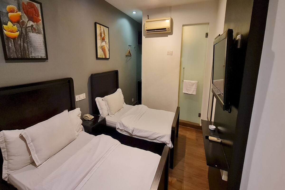Habitación de hotel con dos camas, una mesita de noche con lámpara, aire acondicionado, televisor y un cuadro en la pared.
