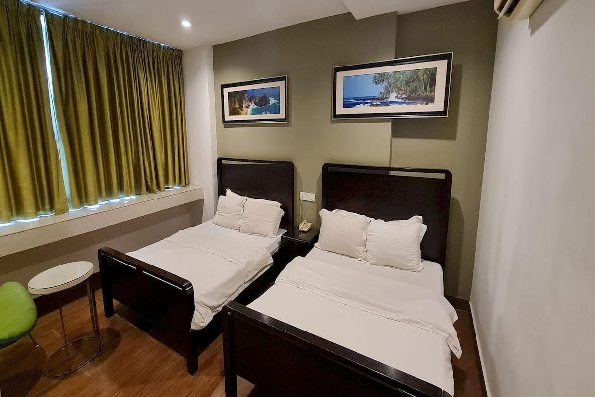Habitación de hotel con dos camas individuales, una mesita redonda pequeña, una silla verde y dos cuadros en la pared.