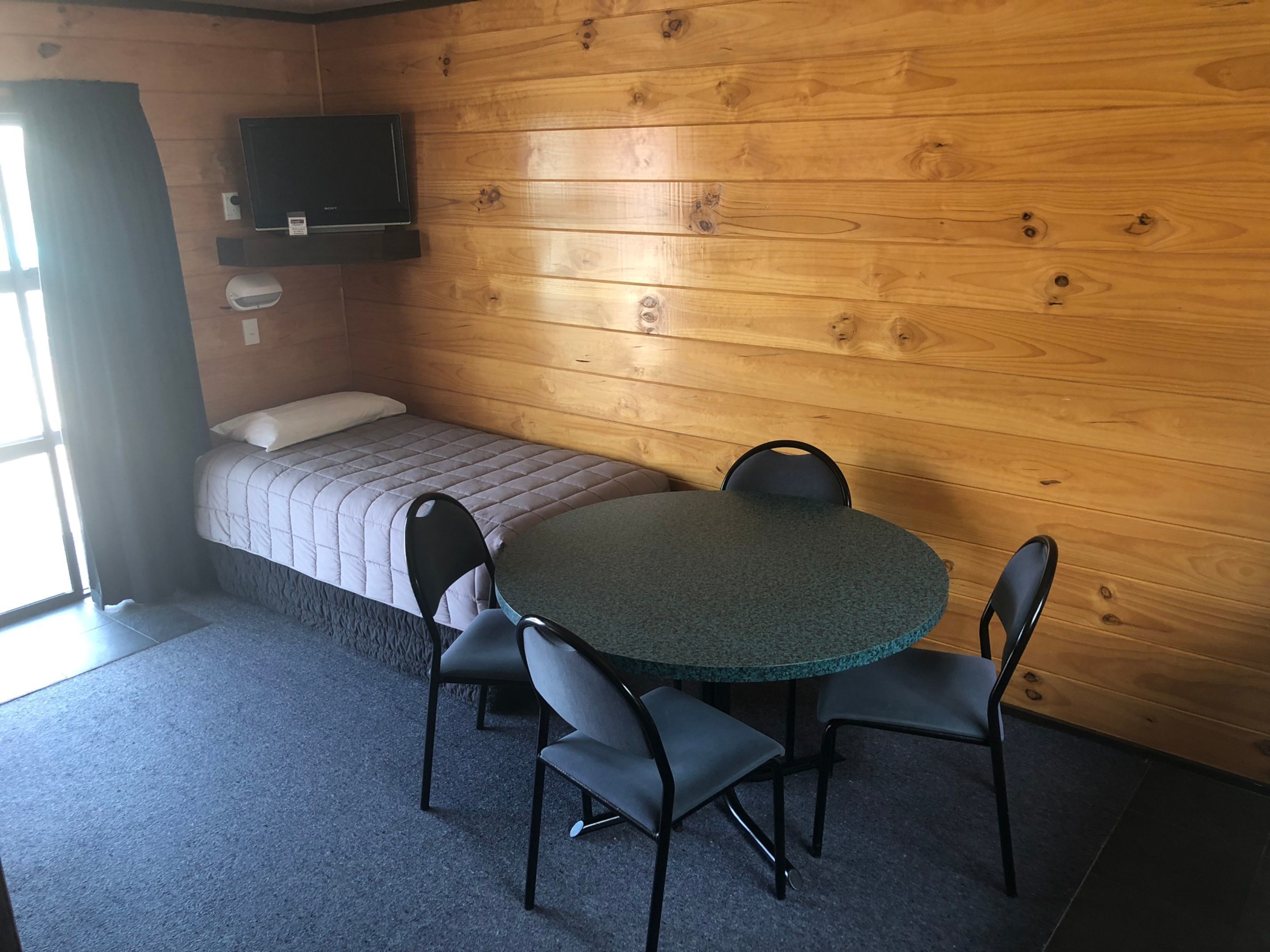 Een kleine kamer met een bed, een ronde tafel met vier stoelen, een aan de muur bevestigde televisie en een raam met gordijnen.