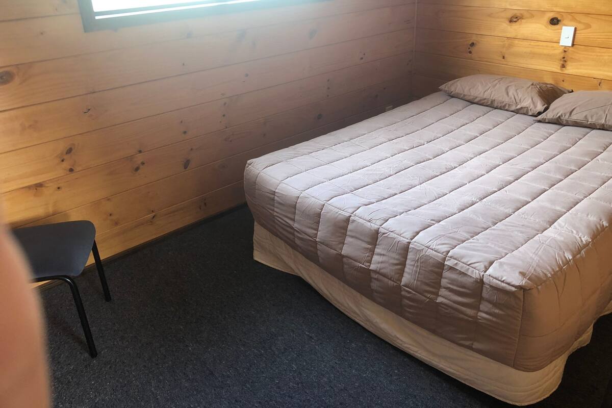 Ein Einzelbett mit einer beigen Bettdecke in einem Zimmer mit Holzwänden und einem kleinen Fenster.
