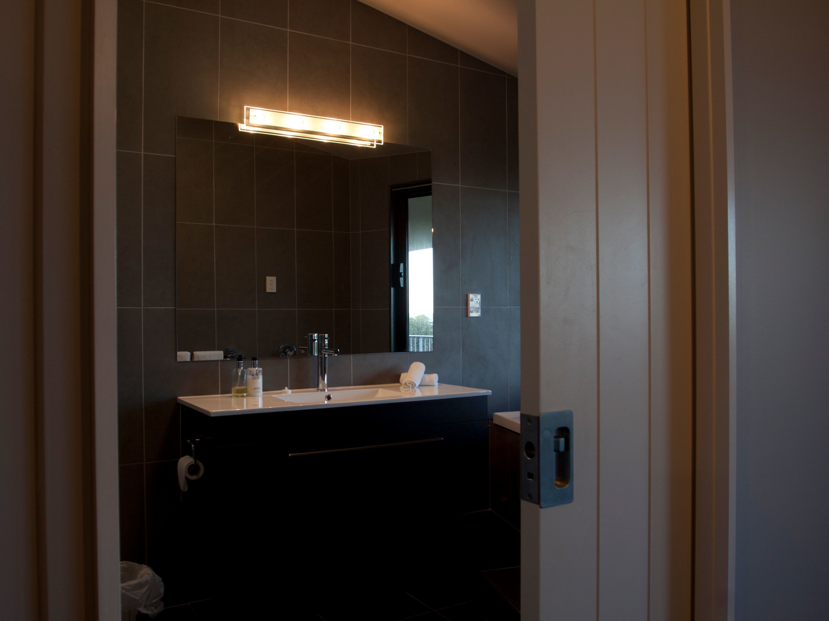 A modern bathroom with a dark tiled wall, a rectangular mirror, a white sink, and a dark vanity.