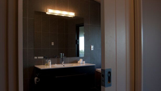 Un baño moderno con una pared de azulejos oscuros, un espejo rectangular, un lavamanos blanco y un mueble de tocador oscuro.