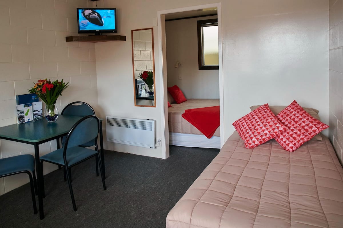 Ein kleiner, gut beleuchteter Raum mit einem Einzelbett, einem Tisch mit Stühlen, einem an der Wand montierten Fernseher und einem Spiegel.