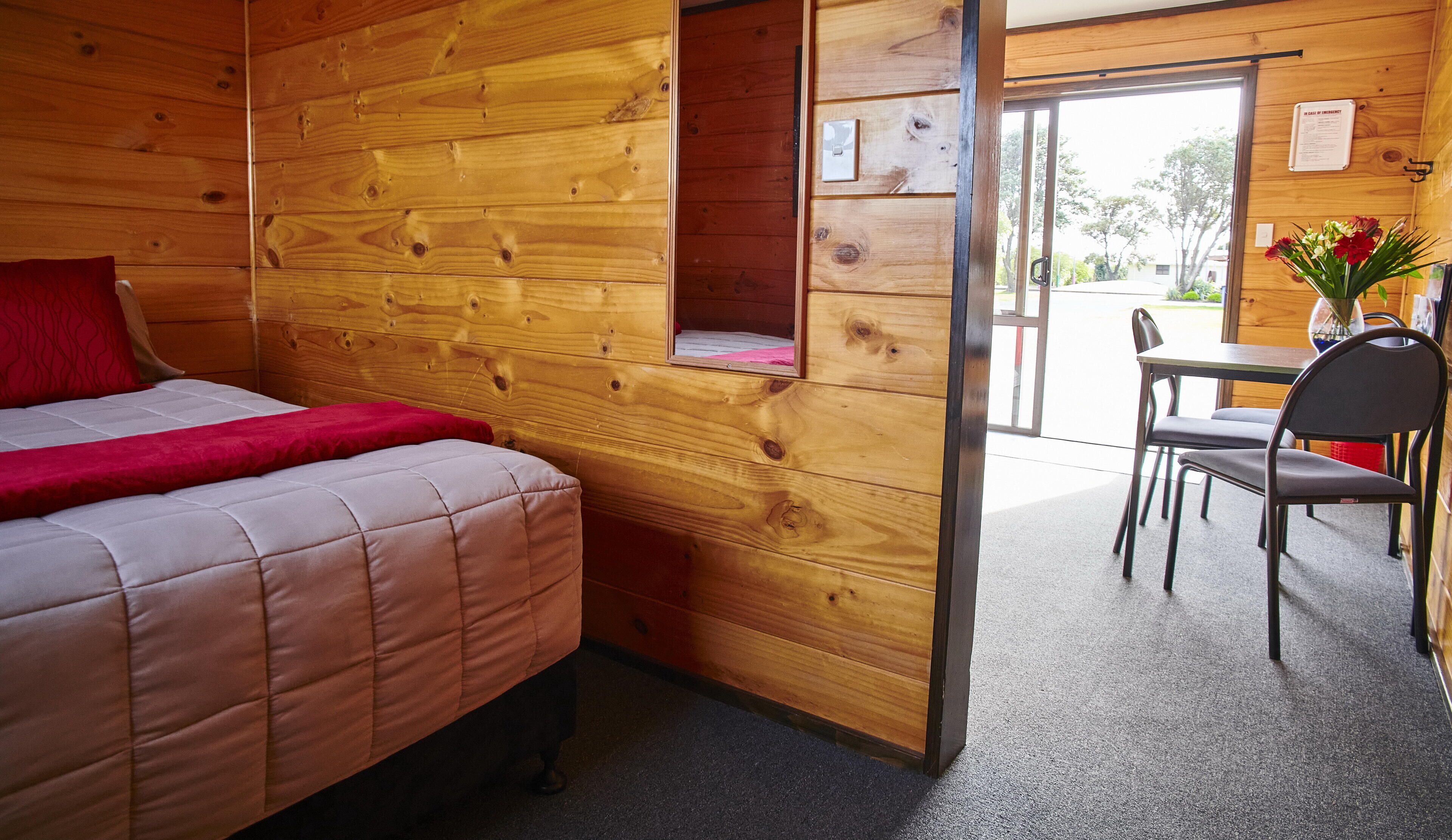 A wooden cabin room with a bed, a small window, a chair, and a table.
