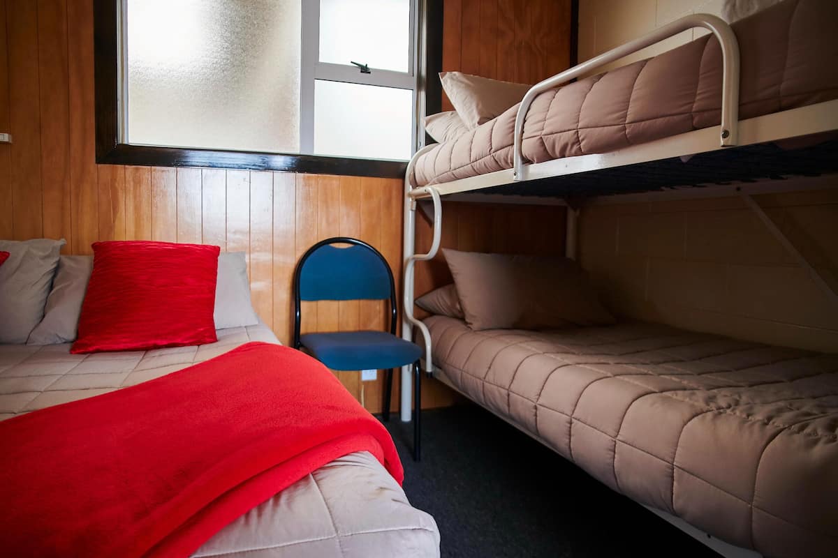 Ein Schlafraum mit Etagenbetten, eine rote Decke auf dem Unterbett, ein blauer Stuhl und ein Fenster mit Jalousien.