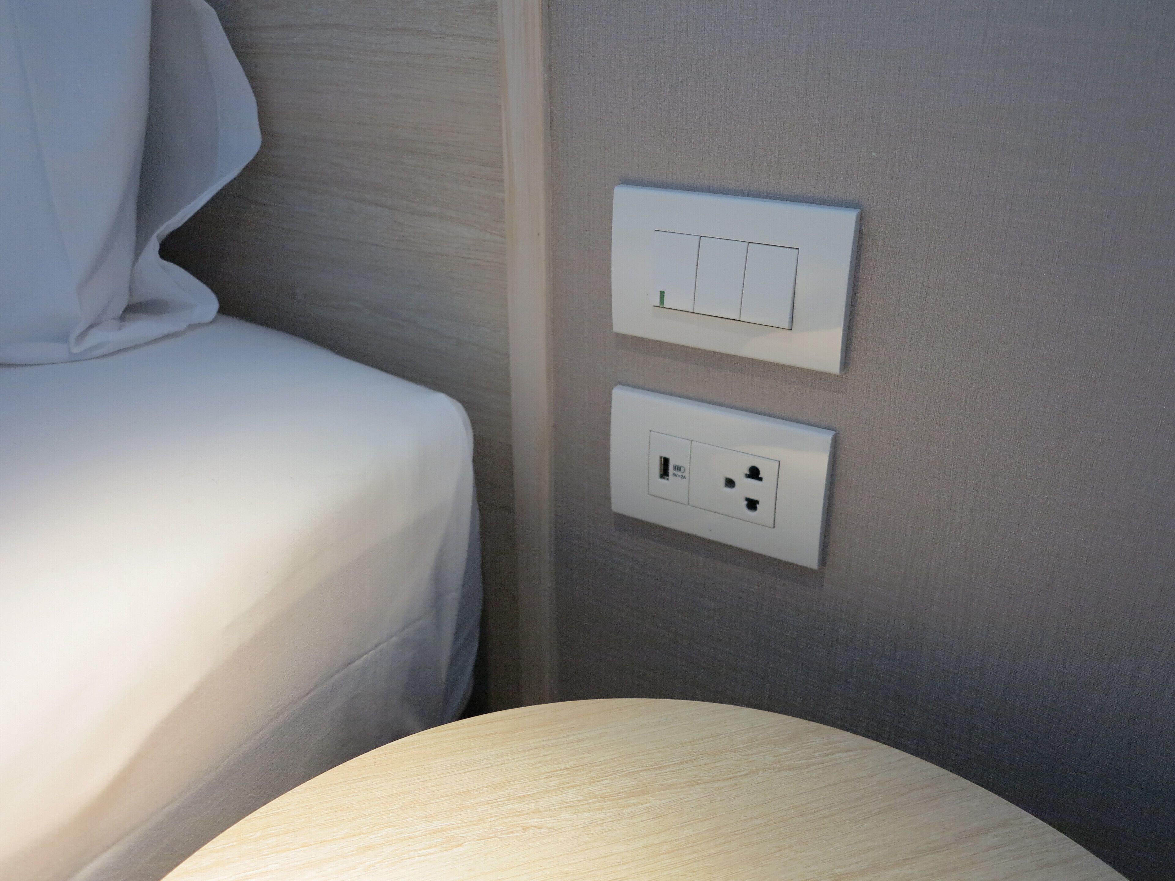 A neatly made bed with a white sheet, a wall-mounted light switch, and a power outlet.
