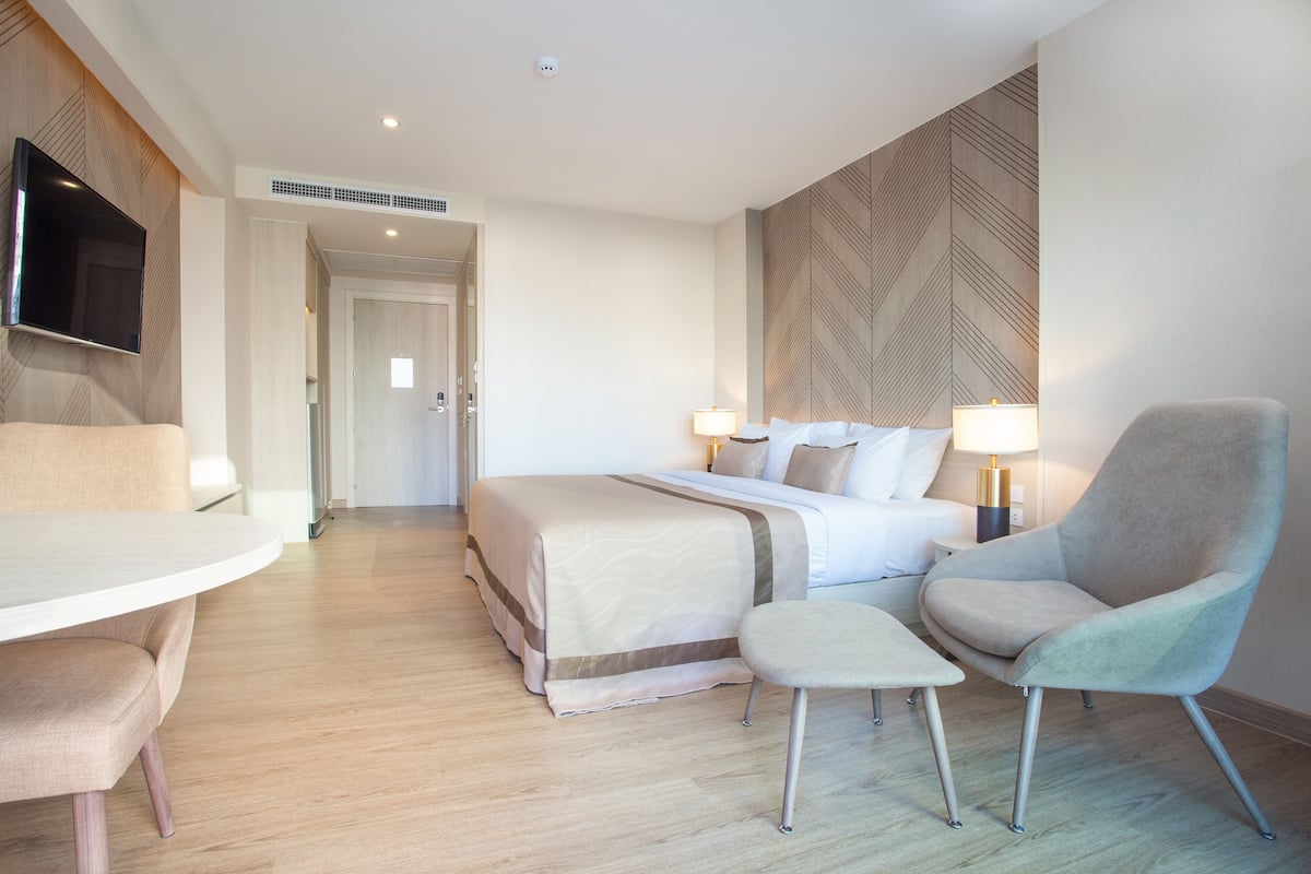 A hotel room with a bed, a desk, a chair, a TV mounted on the wall, and a lamp on the bedside table.