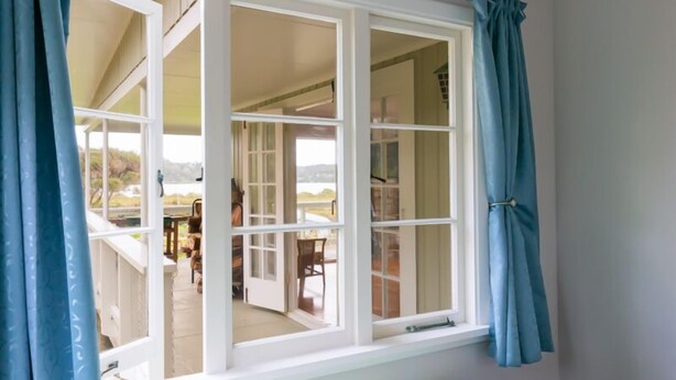 A room with a large window, a bed with striped bedding, and a view of an outdoor patio area. - Russell - Orongo Bay Holiday Park (Russell)
