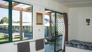 A room with a large sliding glass door leading to an outdoor area, a bed with patterned bedding, and a small table with a chair. - Russell - Orongo Bay Holiday Park (Russell)