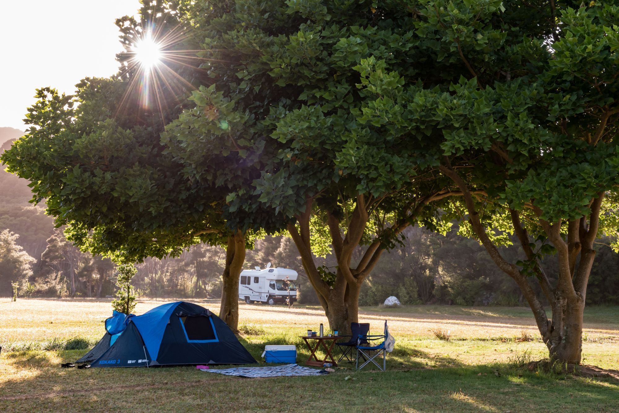 A camping site with a blue tent, a table with chairs, and a white RV in the background.