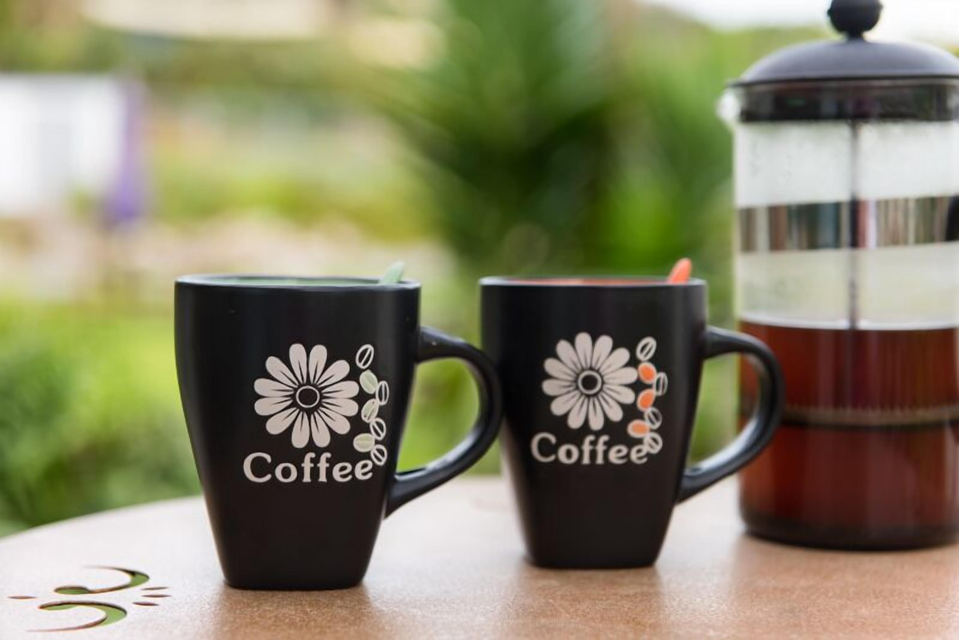 Two black coffee mugs with floral designs and the word "Coffee" on a table with a French press in the background.