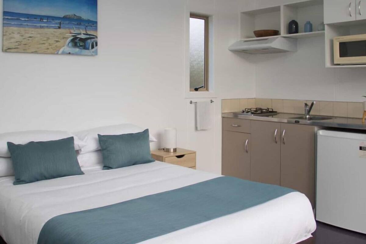 A bedroom with a bed, a nightstand, a painting of a beach scene, a kitchenette with a stove, microwave, and cabinets.
