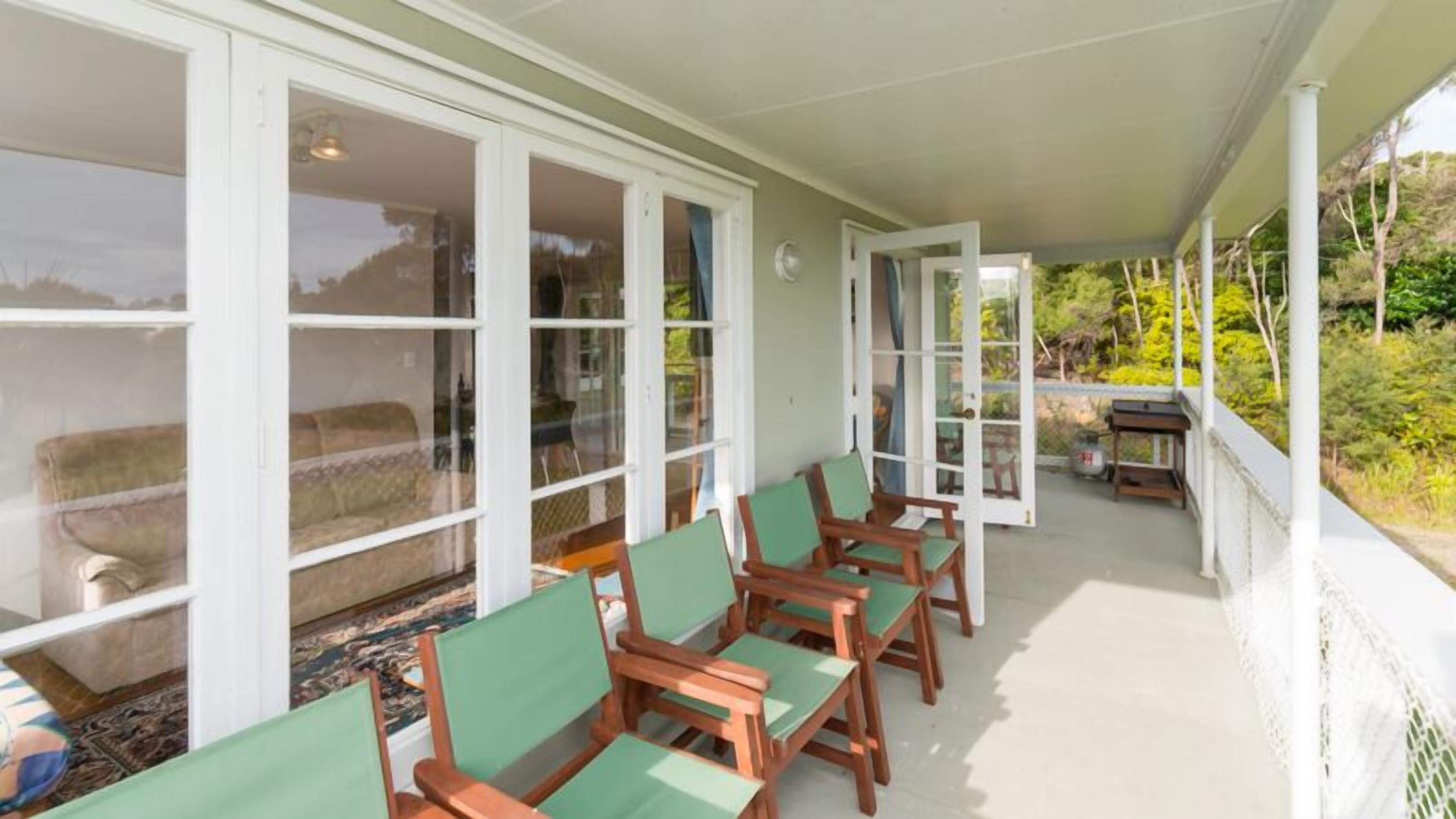 A covered porch with wooden chairs and a table, large windows, and a view of greenery.