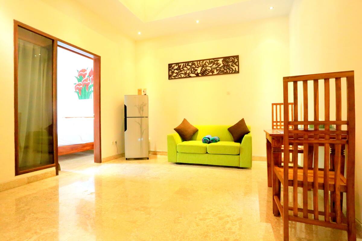 A living room with a green sofa, a wooden chair, a refrigerator, and a decorative wall piece.