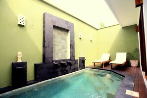 A swimming pool with a stone wall and a window, two lounge chairs, and a small statue. - Jas Green Villas (Seminyak)