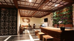 Un área de recepción con un mostrador de madera, varios taburetes de madera, una pared decorativa con plantas, un piso con diseño y un techo con vigas de madera e iluminación empotrada.