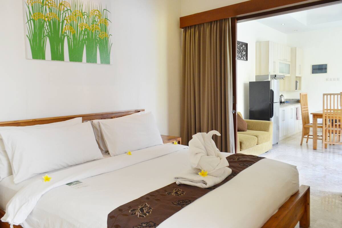 A hotel room with a neatly made bed, a dining area, and a kitchen visible through an open door.