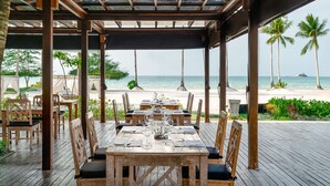 Un espace de restauration en plein air, avec des tables et des chaises en bois, offrant une vue sur une plage bordée de palmiers et sur l’océan.
