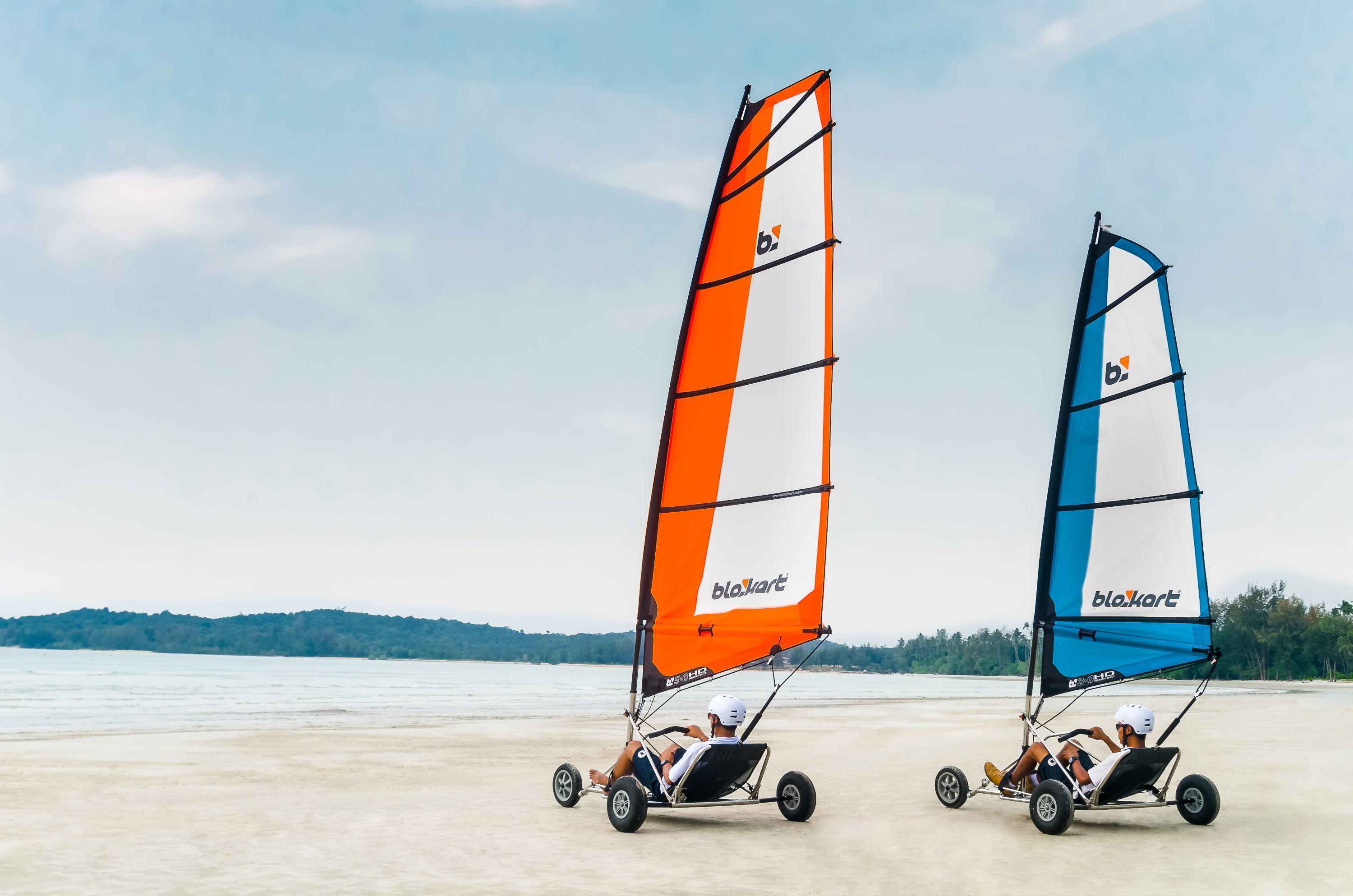 Twee personen in een strandbuggy met zeil, op een zandstrand met water en heuvels op de achtergrond.