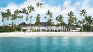 Un hôtel situé en bord de mer, avec une façade blanche, des palmiers et une plage de sable.