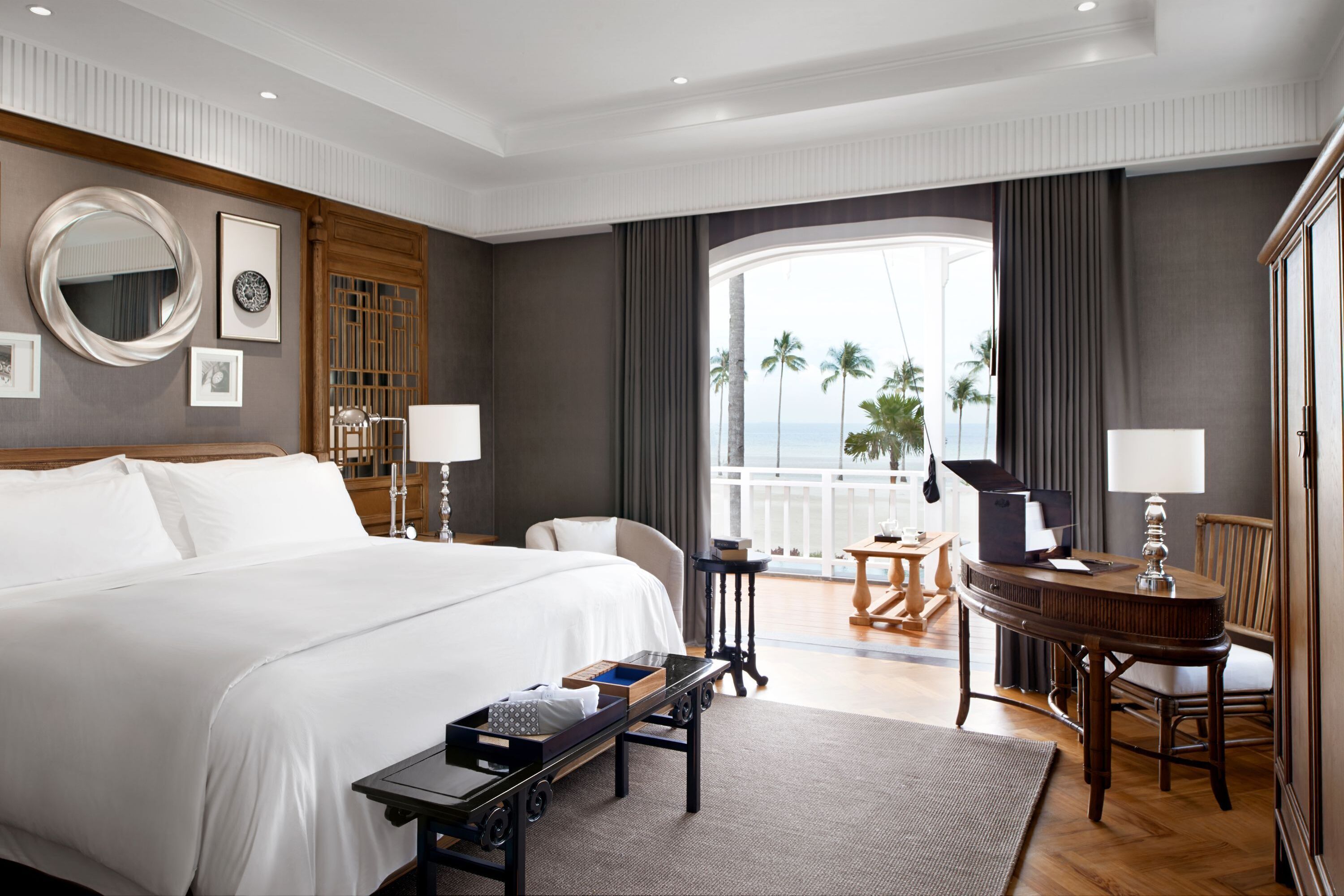 Quarto de hotel com uma cama grande, uma escrivaninha, uma cadeira, um piano e uma varanda com vista para palmeiras.