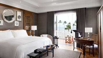 Quarto de hotel com uma cama grande, uma escrivaninha, uma cadeira, um piano e uma varanda com vista para palmeiras.