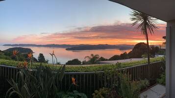 Un balcon offrant une vue sur un coucher de soleil se reflétant sur une étendue d’eau, le tout entouré de plantes tropicales.