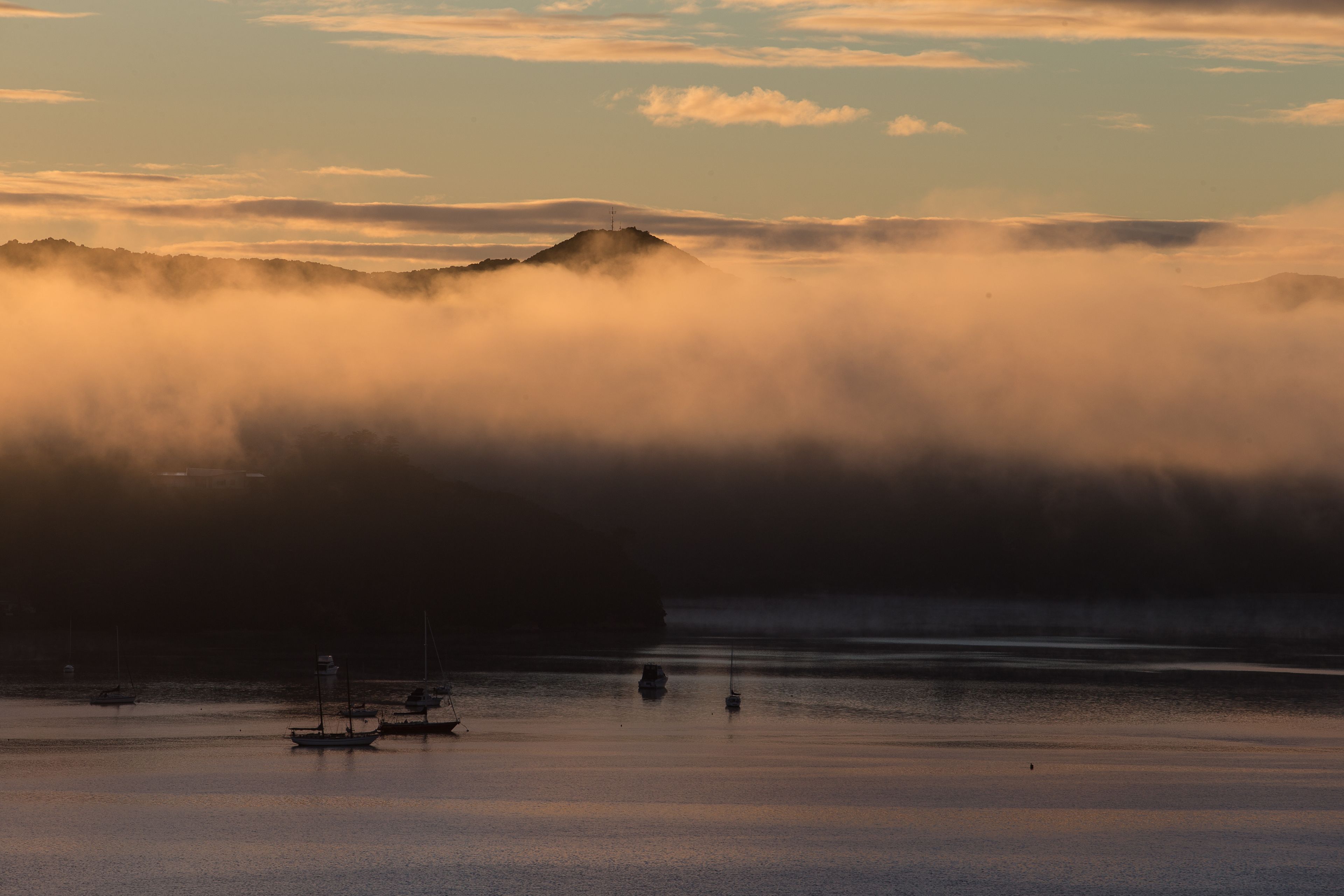 A coastal scene with boats on calm water, a mountain in the background, and a misty atmosphere.