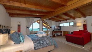 A bedroom with a bed, a red sofa, a TV, and a view of the sea. - Chalet Romantica (Paihia)