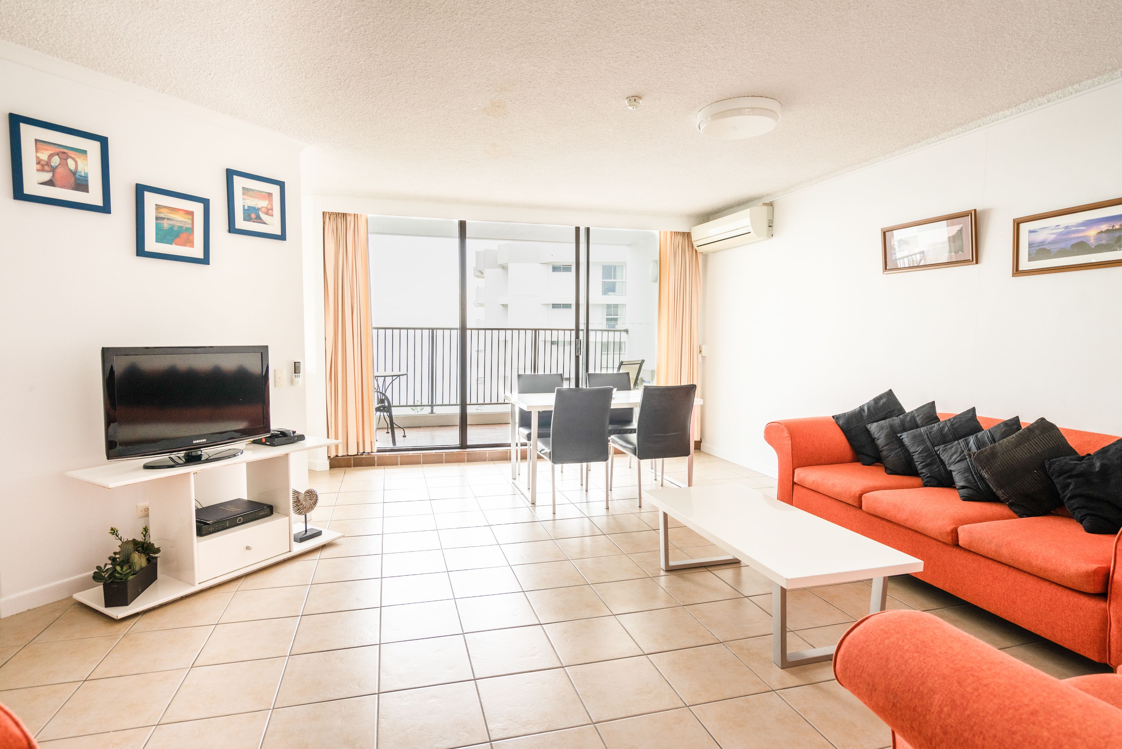 A modern living room with a flat-screen TV, a white coffee table, an orange sofa, and a dining area with chairs.