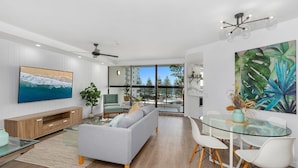 A modern living room with a grey sofa, a glass coffee table, and a TV mounted on the wall. There is a balcony with a view of trees and buildings. - Horizons Holiday Apartments (Burleigh Heads)