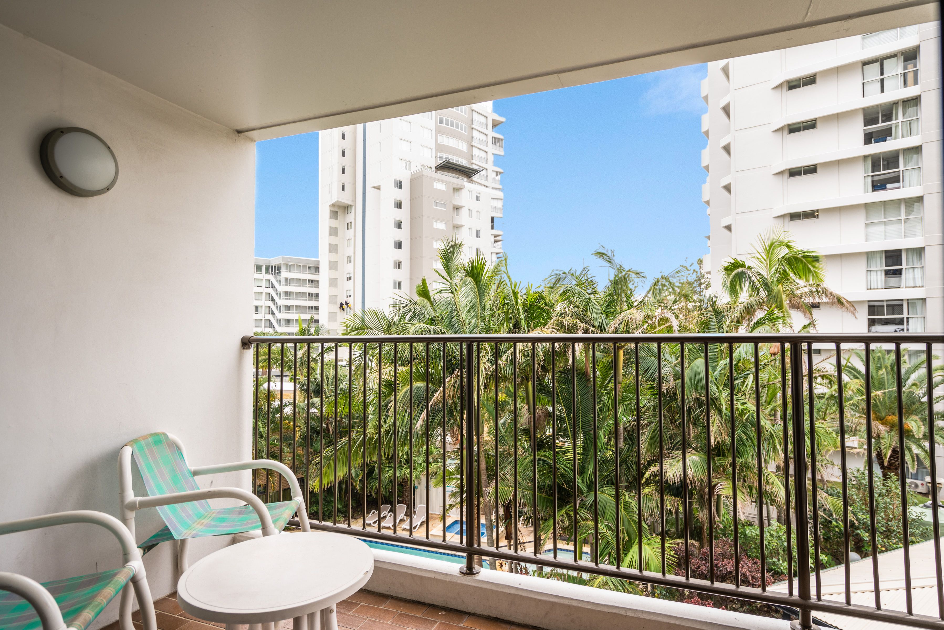 Un balcón con una mesa y sillas blancas, vista a una piscina y edificios altos al fondo.