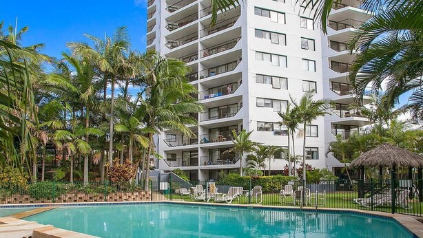 A modern hotel with a swimming pool, surrounded by palm trees and a thatched-roof structure. - Horizons Holiday Apartments (Burleigh Heads)