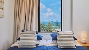 A hotel room with a bed, two pillows, a window with a view of the sea and trees, a lamp, and a painting on the wall. - Horizons Holiday Apartments (Burleigh Heads)