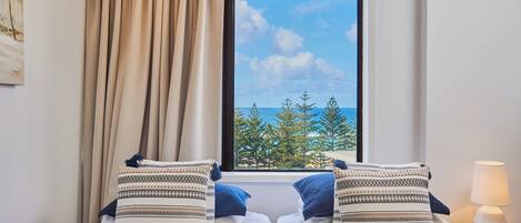 A hotel room with a bed, two pillows, a window with a view of the sea and trees, a lamp, and a painting on the wall.
