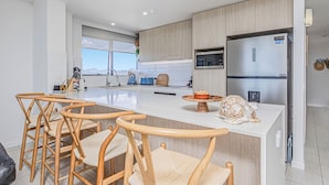 A modern kitchen with a large island, wooden chairs, and a refrigerator. - Horizons Holiday Apartments (Burleigh Heads)