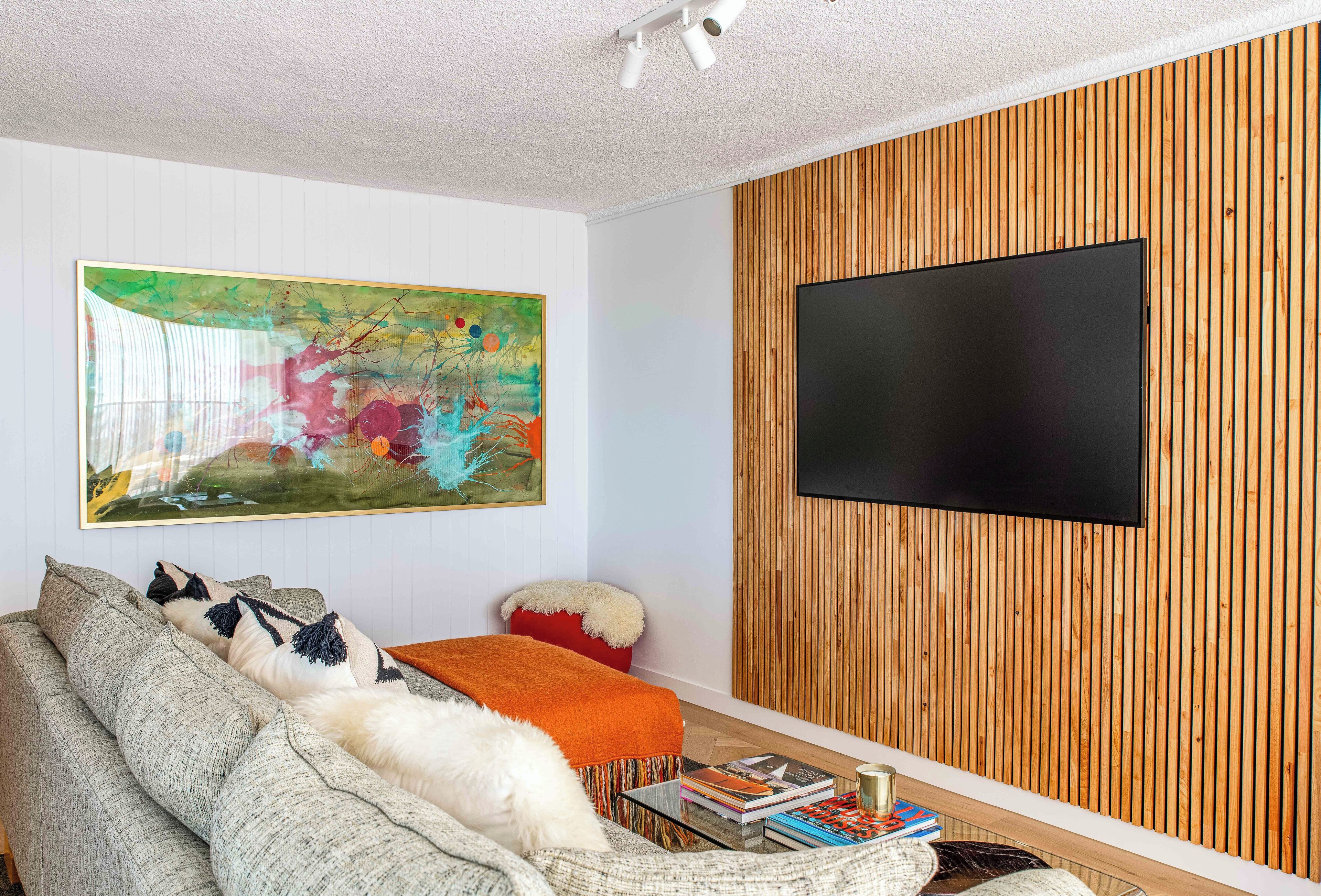 A living room with a sofa, a red ottoman, a flat-screen TV mounted on a wooden wall, and a colorful abstract painting.