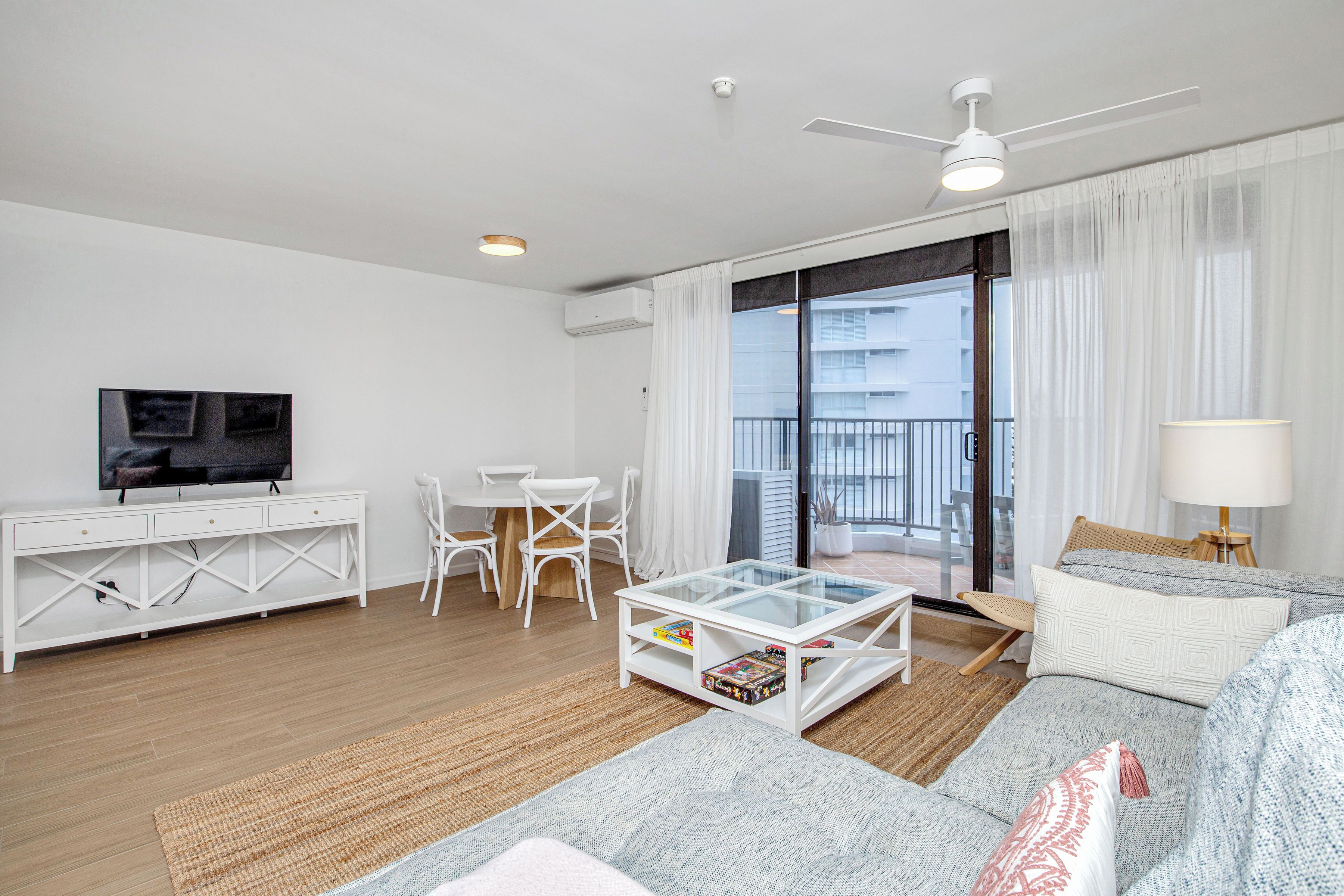 A modern living room with a glass coffee table, white furniture, a flat-screen TV, and a balcony with a view.
