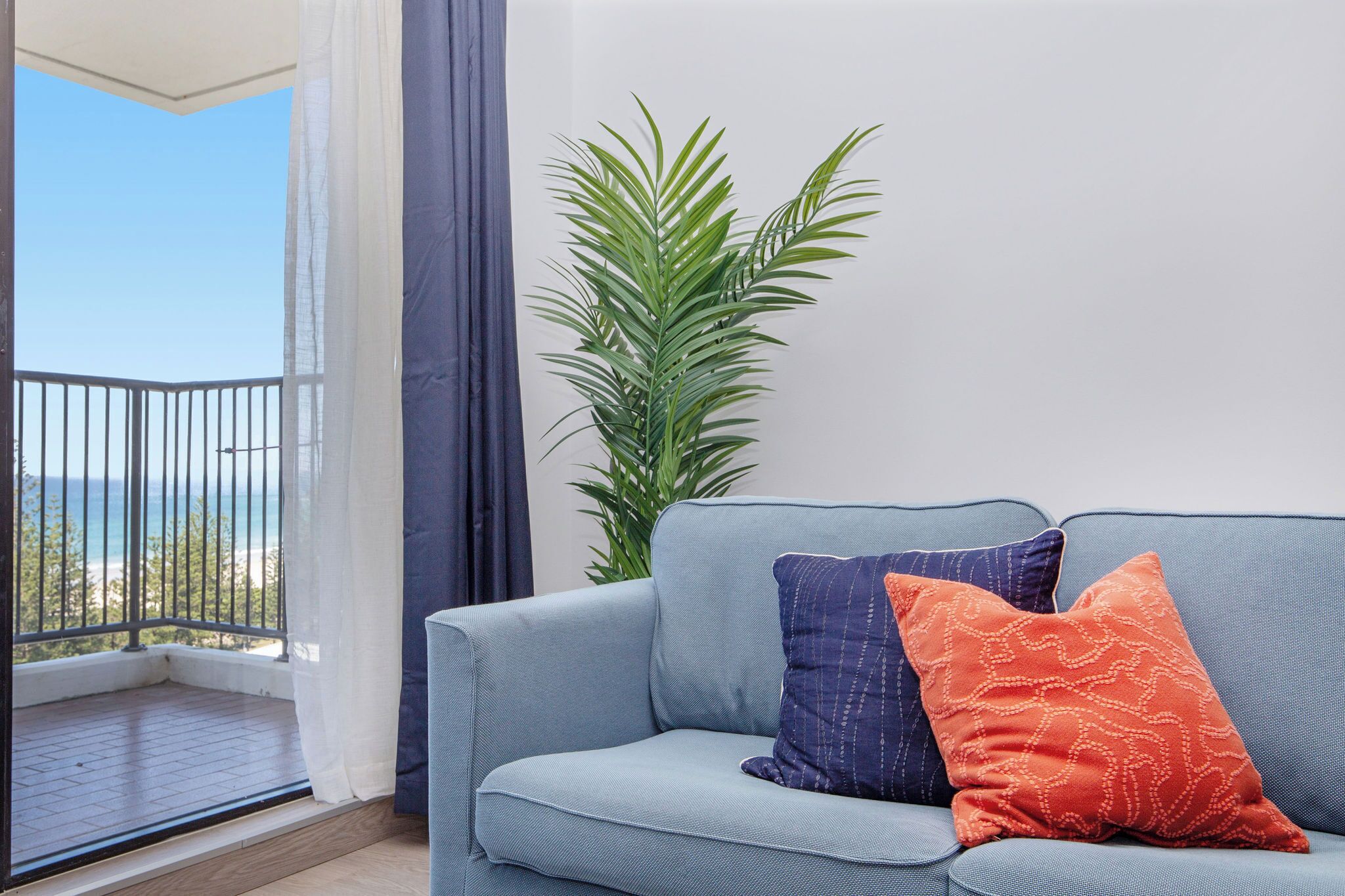 A light blue sofa with two patterned cushions, a potted plant, and a balcony with a view of the sea.