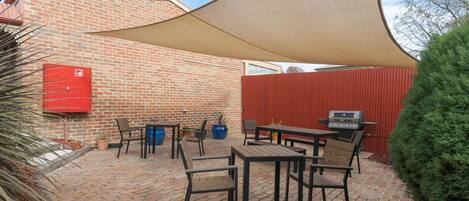Un patio con piso de ladrillo, mobiliario exterior, una parrilla y una vela de sombra grande.