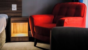 A hotel room with a wooden headboard, a red armchair, a bedside table with a lamp, and a window with a blue curtain. - Yarra Valley Motel (Lilydale)