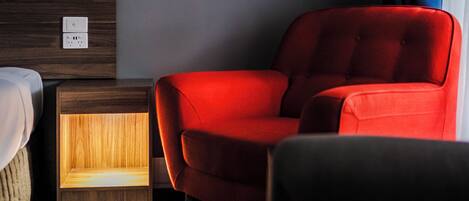 A hotel room with a wooden headboard, a red armchair, a bedside table with a lamp, and a window with a blue curtain.