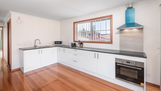 A modern kitchen with white cabinets, a black countertop, a built-in oven, and a window with blinds. - Yarra Valley Motel (Lilydale)