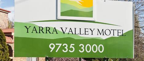 A Yarra Valley Motel sign with a sun and landscape graphic, surrounded by well-maintained hedges and a clear blue sky.
