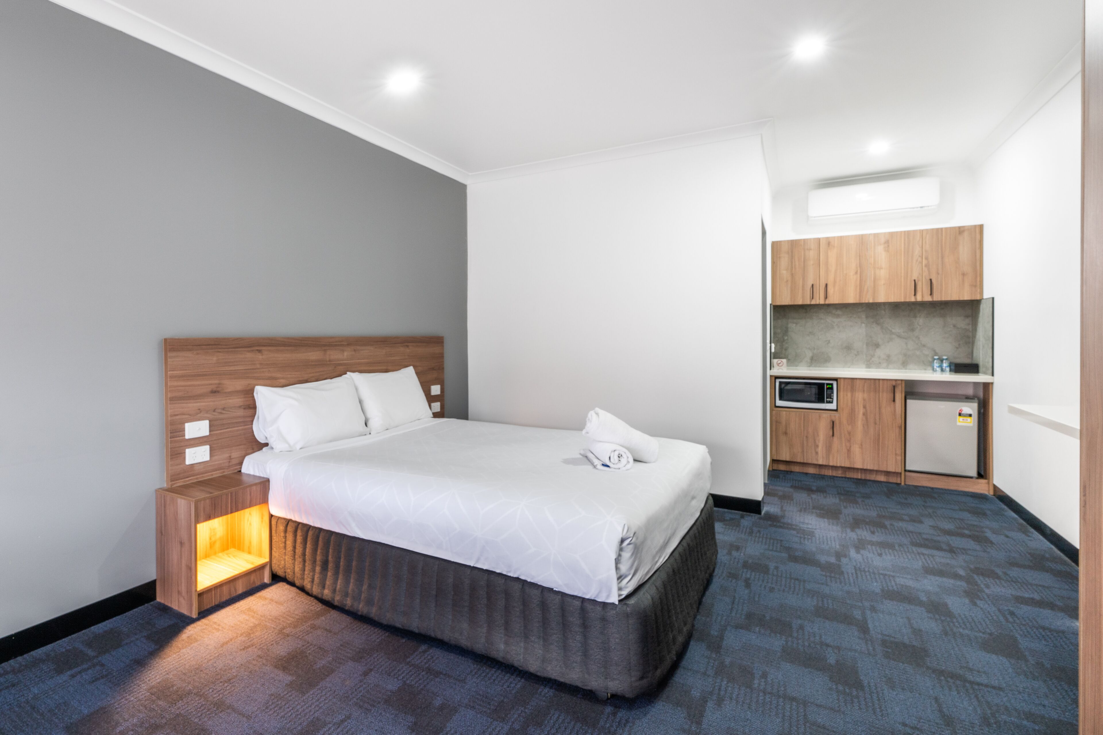 Quarto de hotel moderno com uma cama bem arrumada, uma pequena mesa de cabeceira com abajur, área de cozinha integrada com micro-ondas e geladeira, e uma grande janela que permite entrada de luz natural.