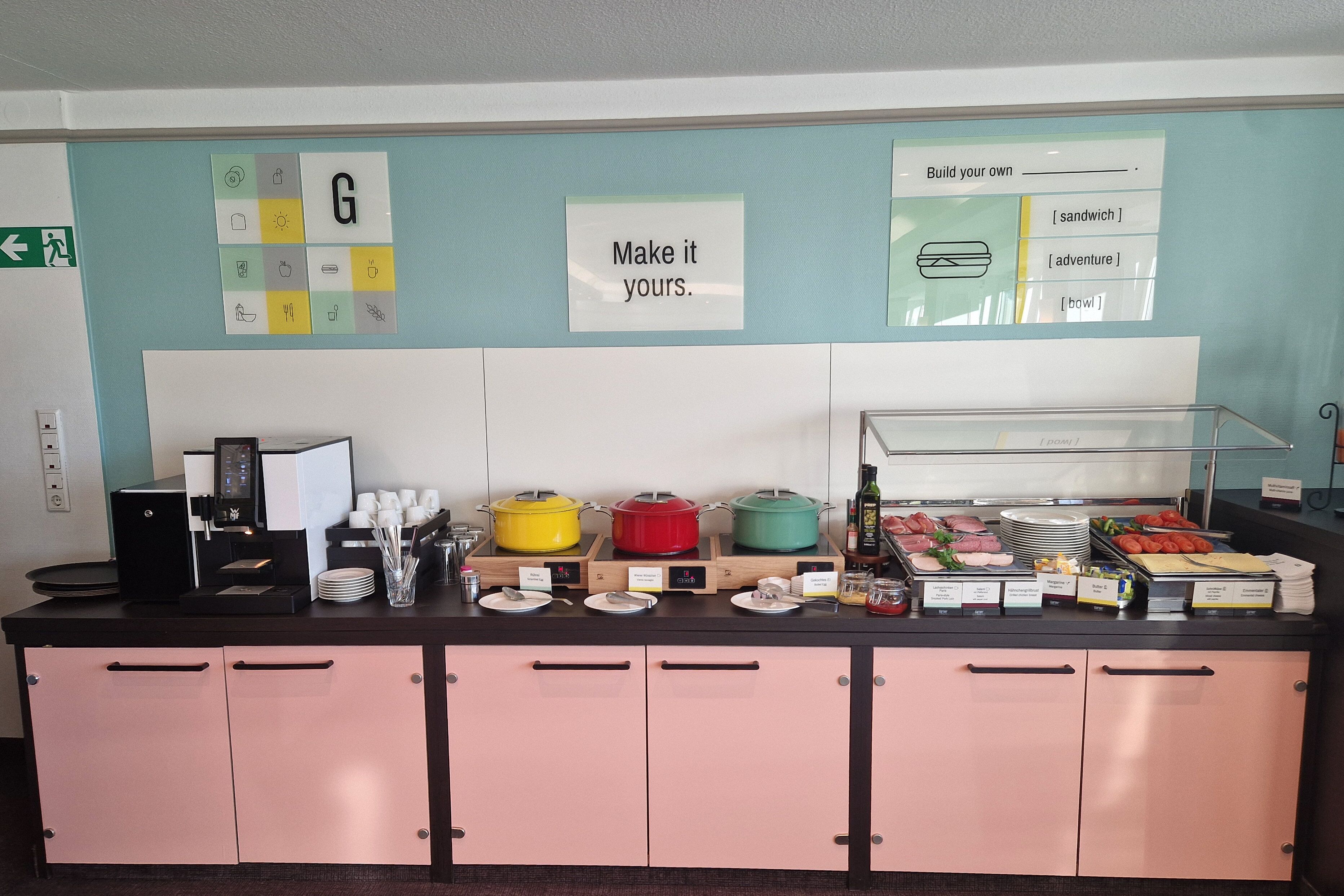 Mostrador de buffet con una máquina de café, diferentes opciones de comida y un letrero que dice 'Make it yours'.