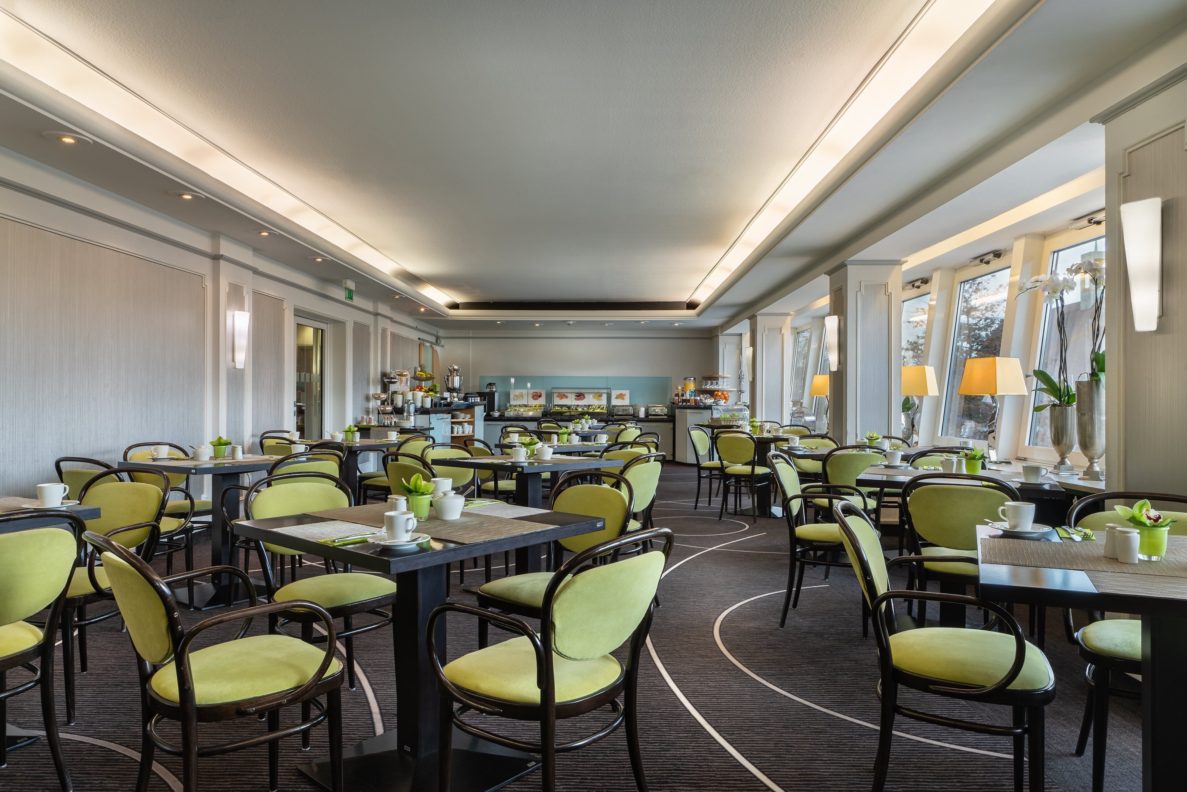 A hotel dining area with multiple tables and chairs, a kitchen in the background, and large windows.