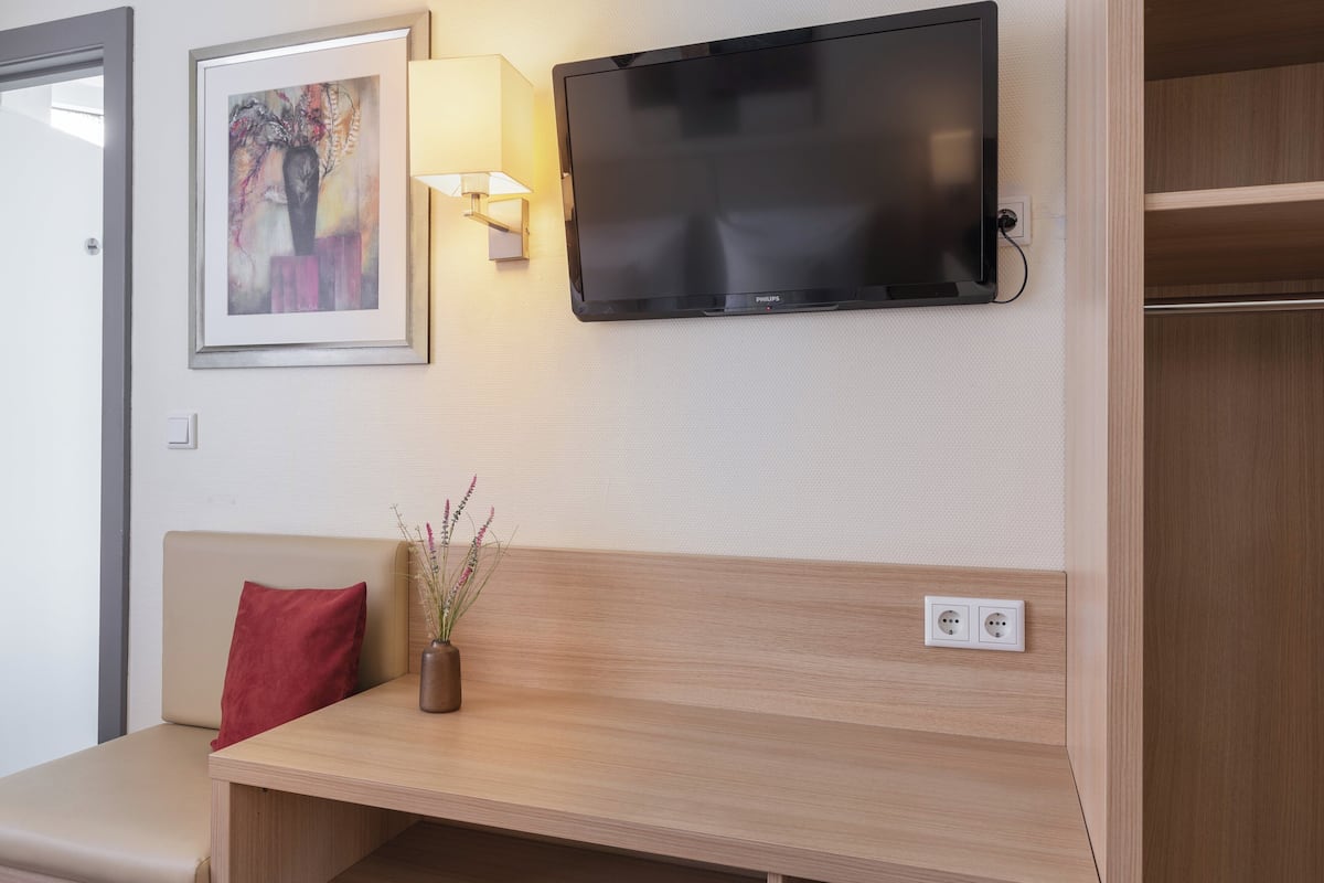 A hotel room with a flat-screen TV mounted on the wall, a bedside table with a vase of flowers, a red pillow, and a wooden bench.