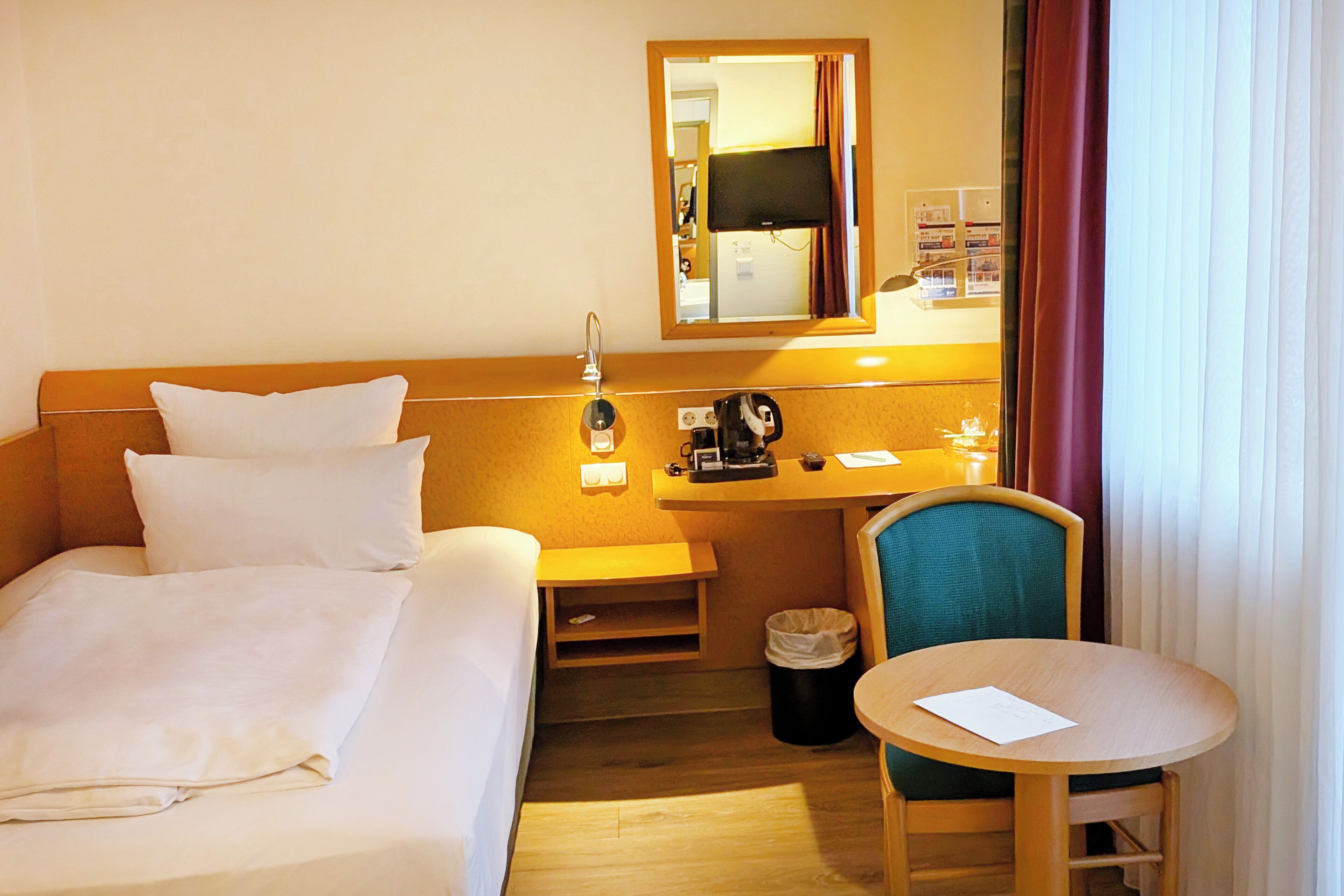 A hotel room with a bed, a desk, a chair, a trash bin, and a small table.